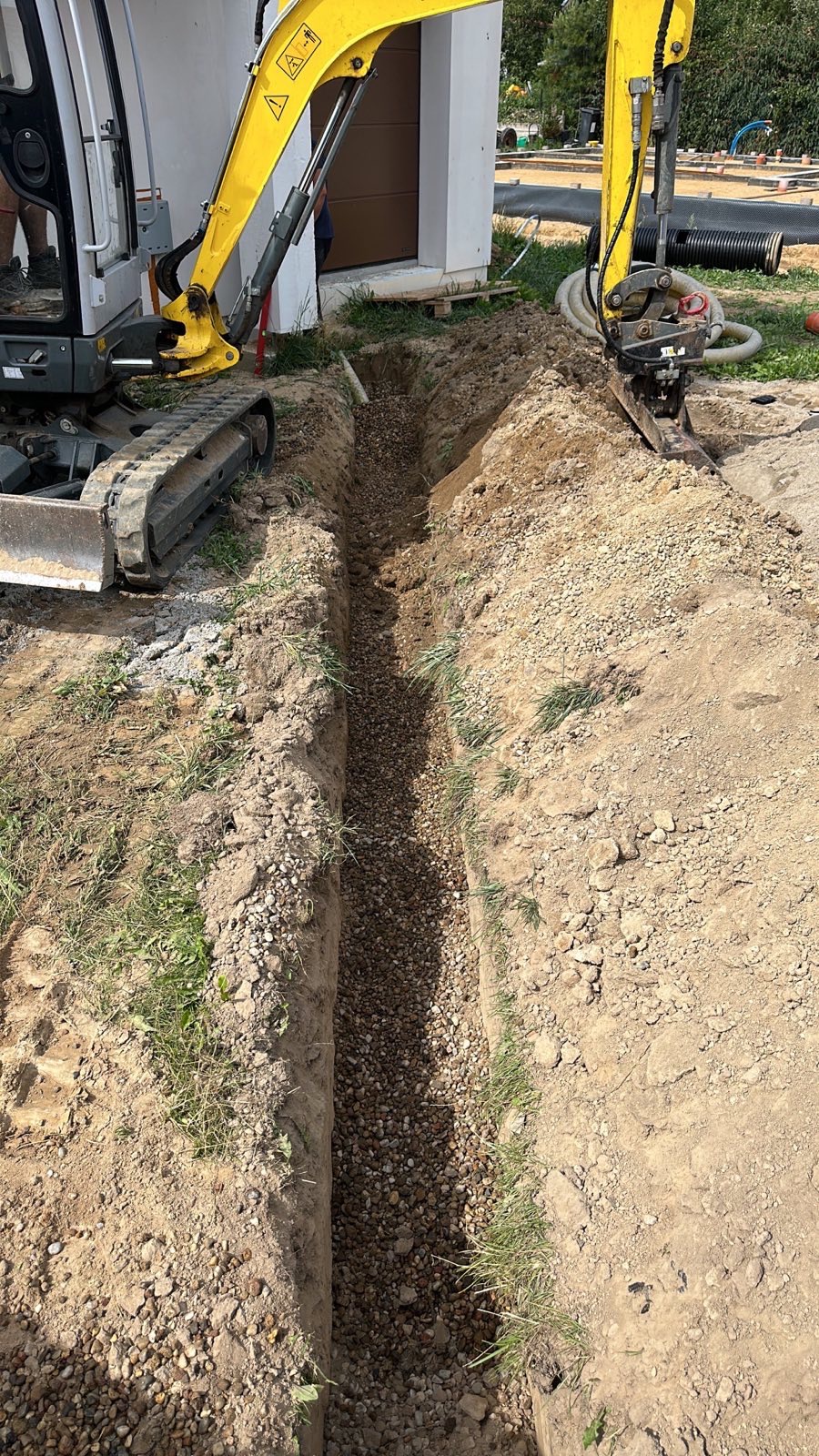 Długi, wąski wykop wypełniony drobnym żwirem, obok stoją dwa żółte mini-koparki, w tle budynek z brązową bramą garażową.