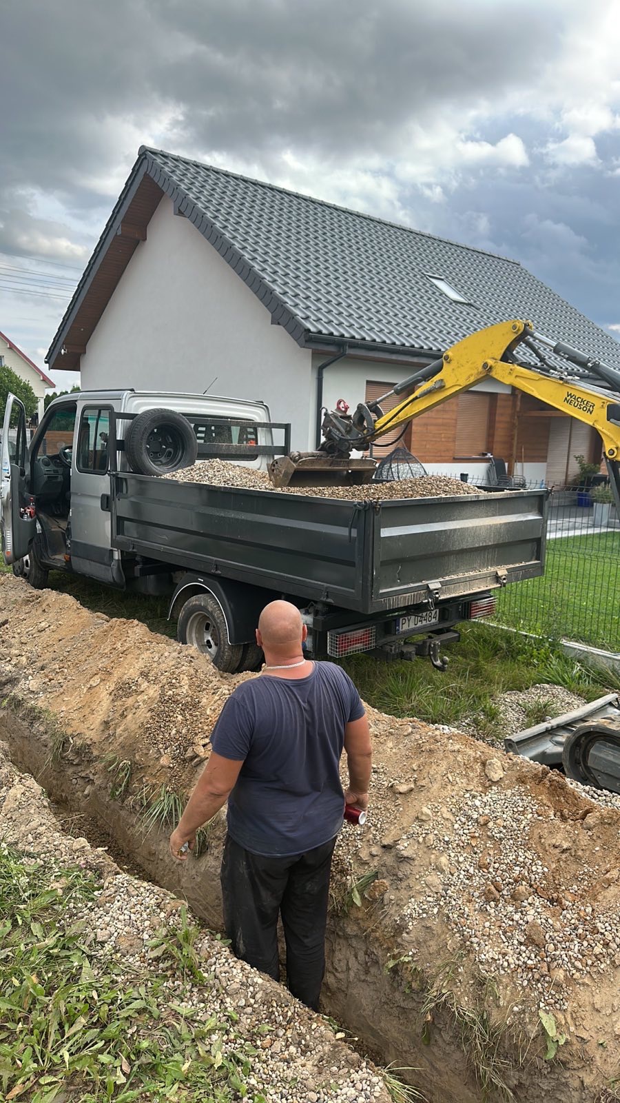 Wykopany rów z podsypką żwirową, srebrny samochód dostawczy z wywrotką wypełnioną żwirem i mini koparka w tle, pracownik nadzoruje prace.