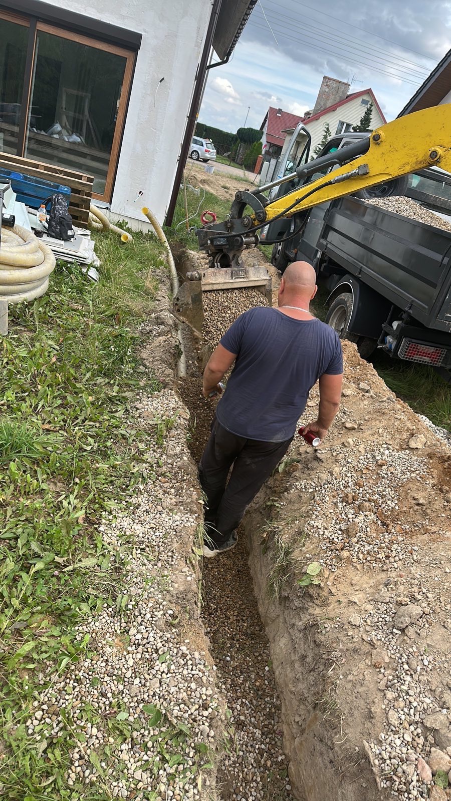 Robotnik w wykopie zasypanym żwirem, obok koparka z przyczepą wypełnioną żwirem, dom w tle.