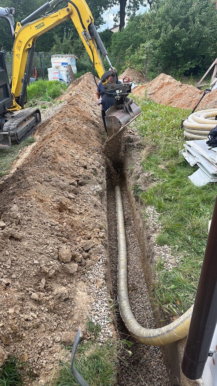 Żółta koparka Wacker Neuson zasypuje wykop z ułożoną rurą drenażową drobnym żwirem, widoczny operator koparki i hałda ziemi w tle.