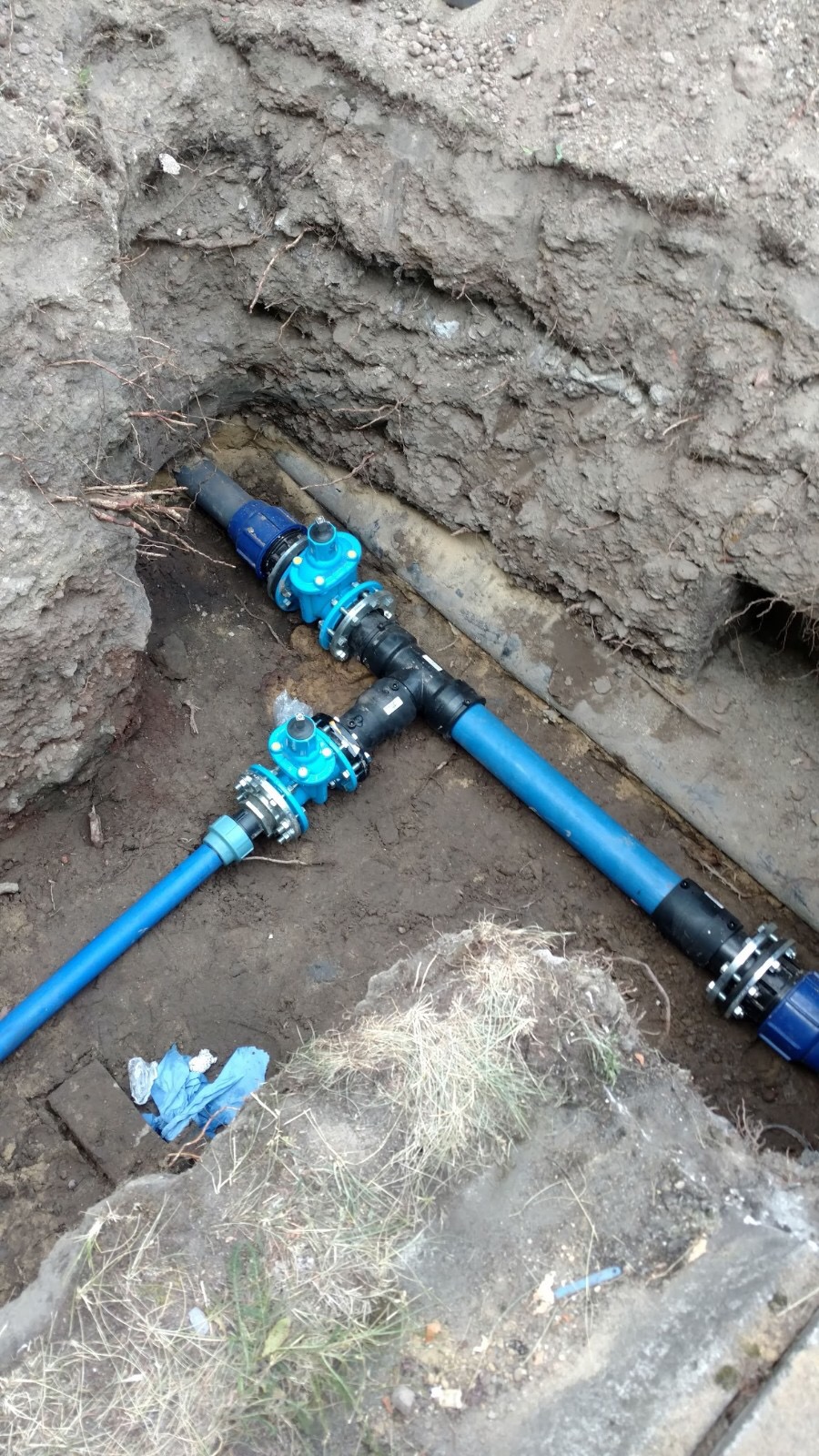 Niebieskie rury wodociągowe z zaworami w wykopie ziemnym, widoczne połączenia skręcane, w tle fragment starej rury.