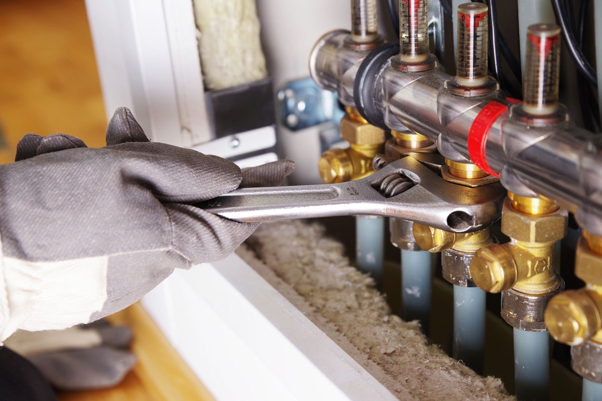 Szczegółowy widok dłoni w szarej rękawicy roboczej, dokręcającej kluczem nastawnym mosiężną złączkę w rozdzielaczu instalacji grzewczej, z widocznymi rurkami i przepływomierzami.