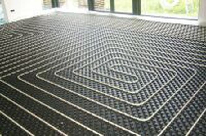 Widok z góry na rozłożone rury ogrzewania podłogowego, białe rurki ułożone w regularnych pętlach na czarnej macie izolacyjnej, przygotowane do zalania wylewką.