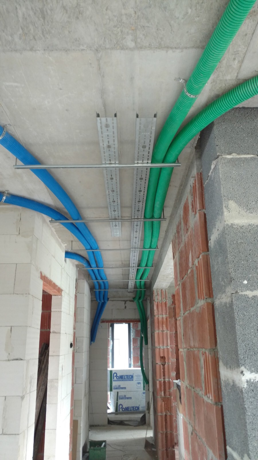 Niebieskie i zielone rury wentylacyjne poprowadzone pod sufitem w surowym stanie, widoczne metalowe korytka montażowe, ściany z białego i czerwonego pustaka.