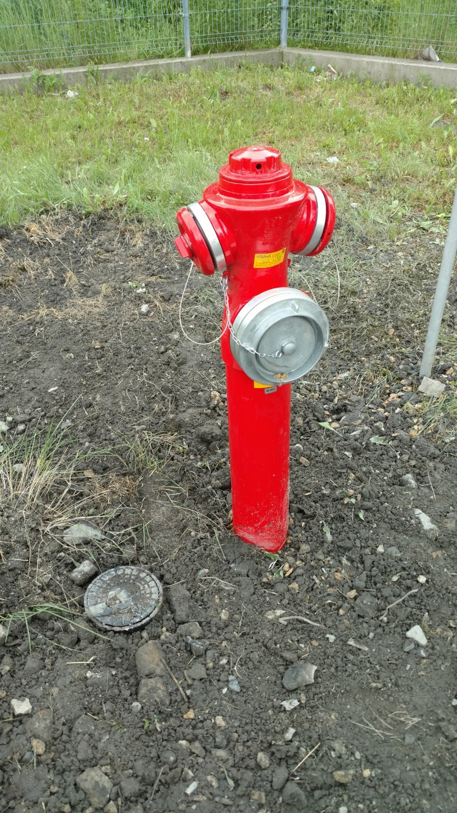Czerwony hydrant naziemny z metalową pokrywą na łańcuszku, stojący na ziemi z widoczną studzienką w tle, otoczony trawą i ogrodzeniem.