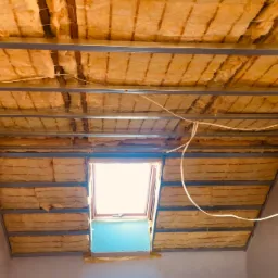 Izolacja poddasza wełną mineralną z widoczną konstrukcją metalową i oknem dachowym w trakcie montażu.