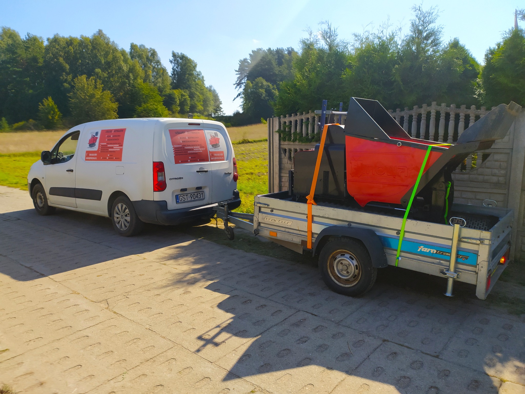 Biały samochód dostawczy Citroen ciągnie przyczepę z czerwono-czarnym rębakiem do gałęzi, zaparkowany na betonowej nawierzchni z zielonym tłem drzew.