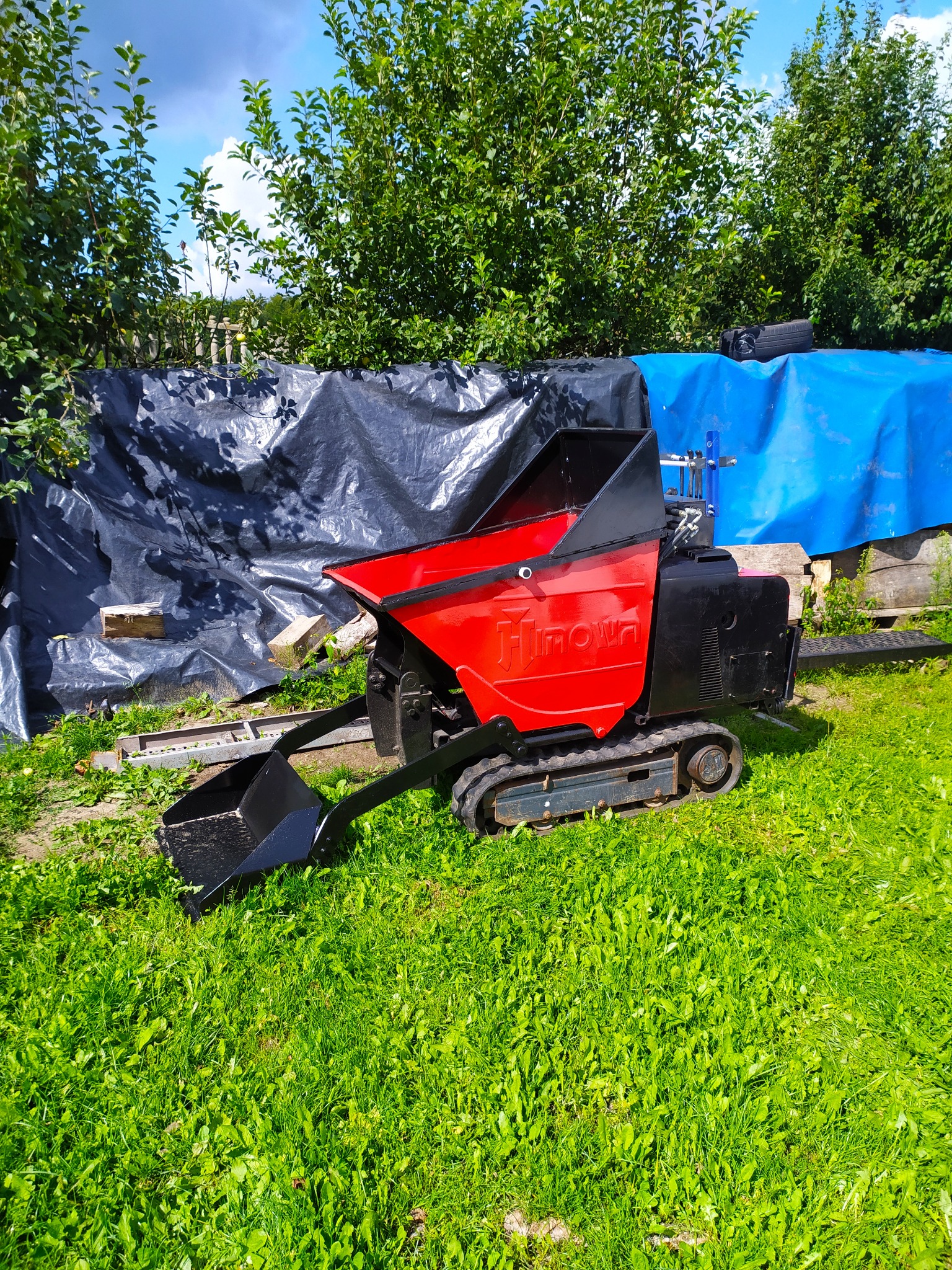 Czerwono-czarny mini dumper gąsienicowy marki Hinowa stoi na trawie, w tle czarna i niebieska plandeka oraz zieleń drzew.