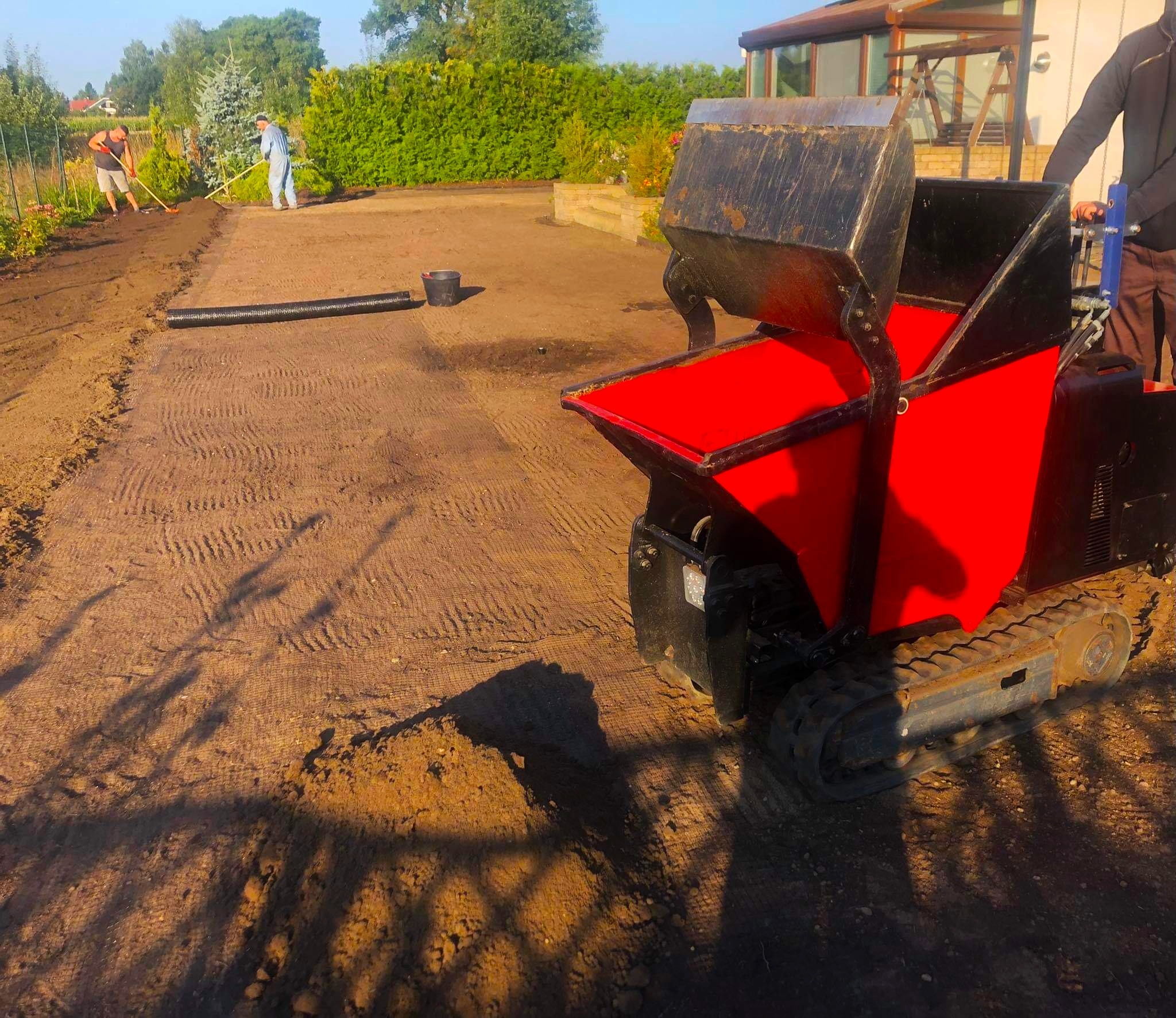 Niwelacja terenu pod trawnik z użyciem minikoparki gąsienicowej z czerwoną skrzynią, widoczni pracownicy z grabiami wyrównujący ziemię w tle, przygotowanie pod trawnik z rolki.