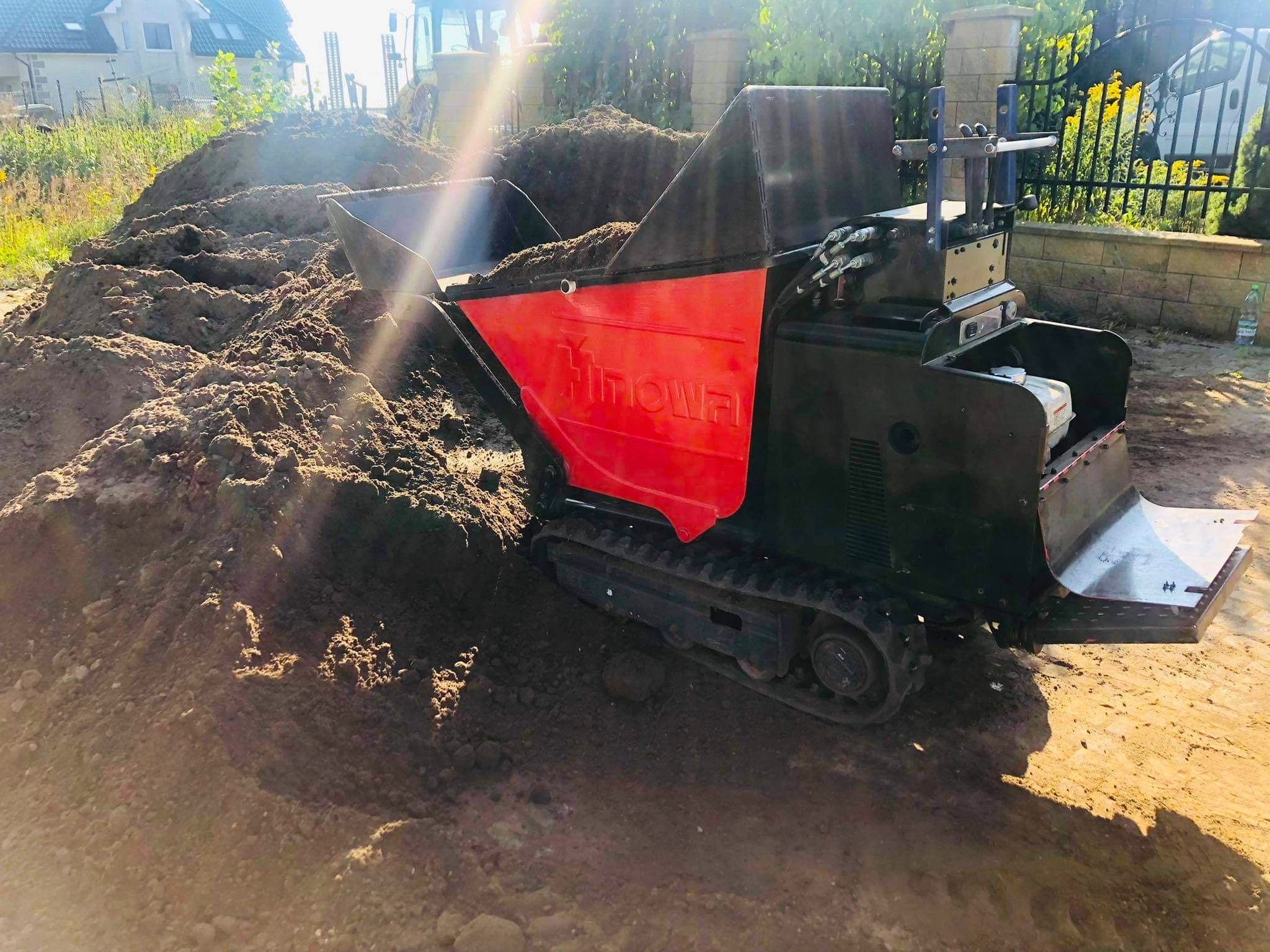 Mini-wywrotka gąsienicowa marki Hinowa, wypełniona ziemią, na placu budowy w słoneczny dzień; w tle dom jednorodzinny i ogrodzenie.