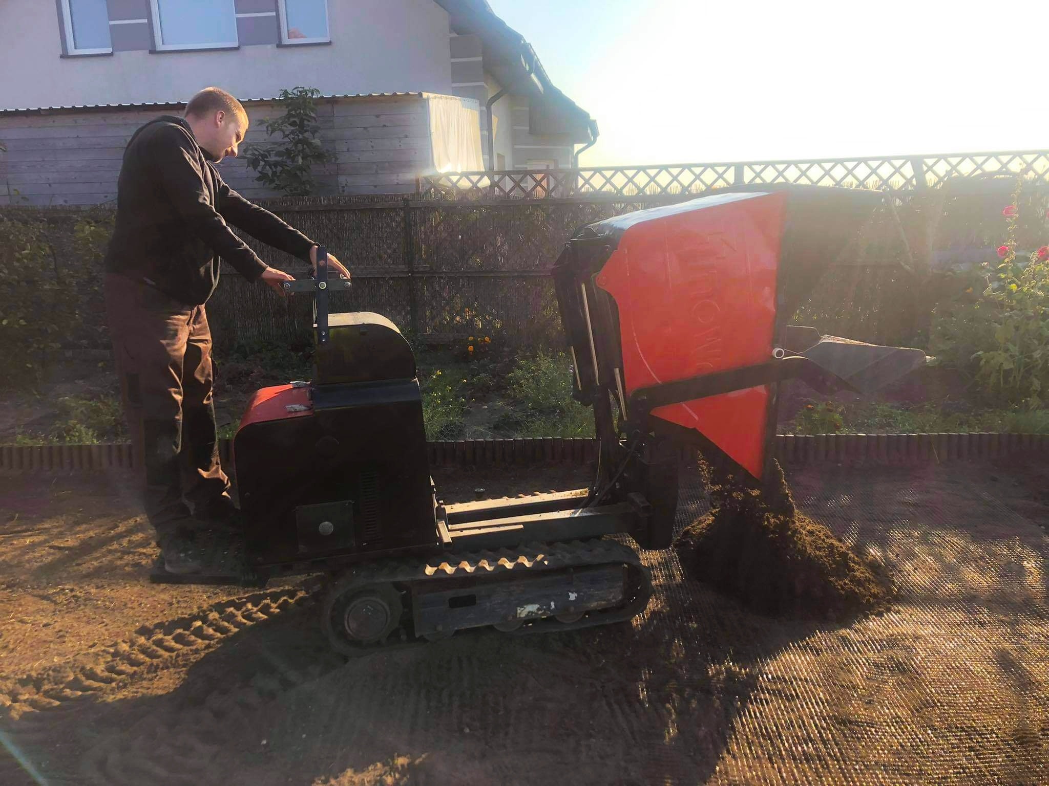 Operator wysypuje ziemię z mini-wywrotki gąsienicowej na siatkę wzmacniającą grunt; w tle ogrodzenie i budynek mieszkalny.