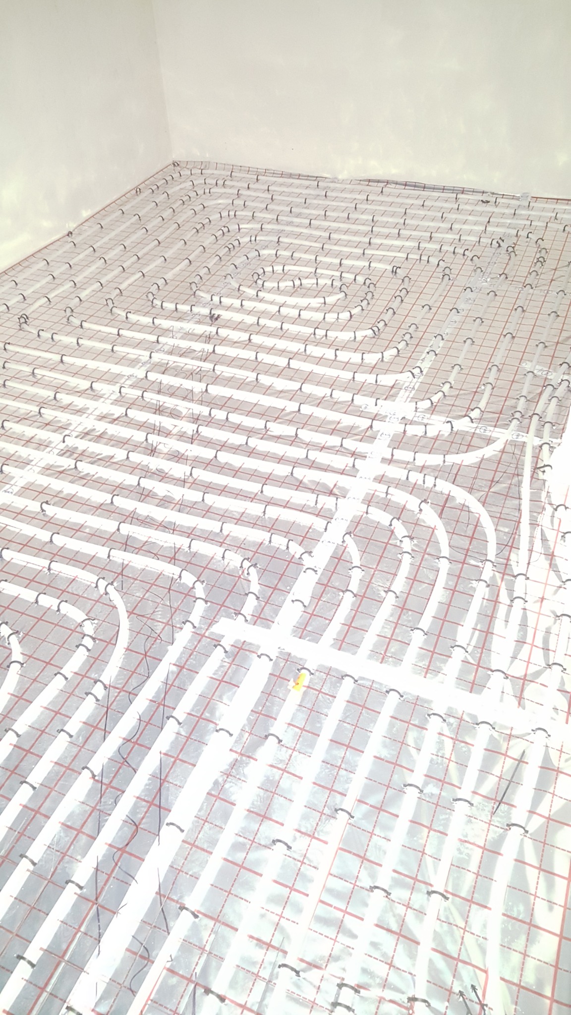 Rozłożona instalacja ogrzewania podłogowego na folii z rastrem, widoczne białe rurki i czarne zaczepy, przygotowane do zalania wylewką.