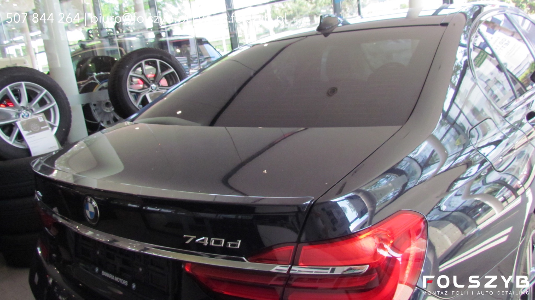Ciemna folia na tylnej szybie czarnego BMW 740d, widoczne logo firmy montującej folie w prawym dolnym rogu, w tle opony i felgi samochodowe.