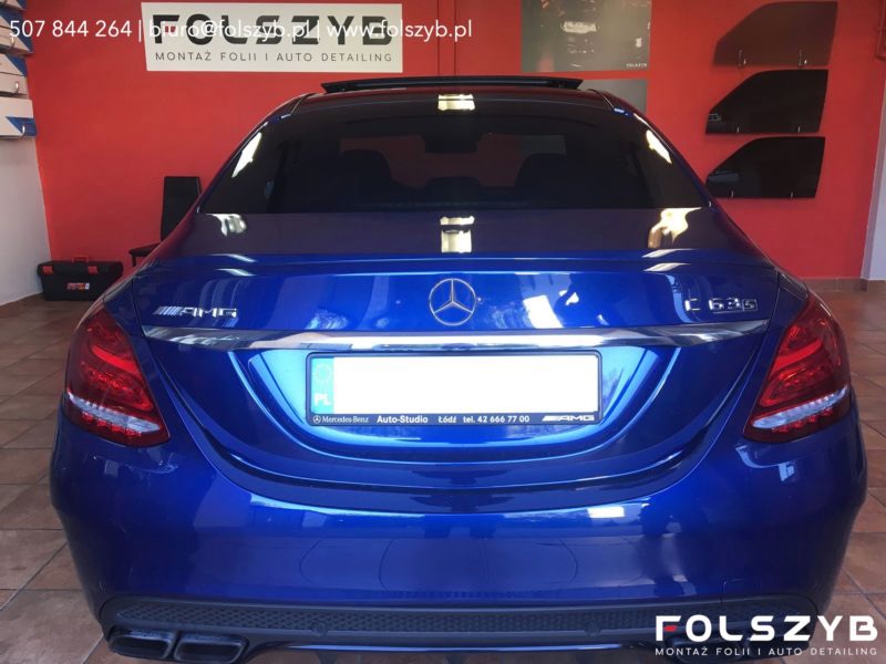 Niebieski Mercedes C63S z przyciemnionymi tylnymi szybami i oknem dachowym, widok z tyłu, z widocznymi próbkami folii na ścianie w tle.