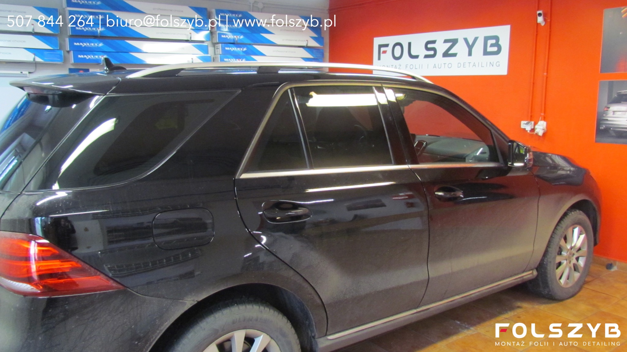 Czarny SUV z przyciemnionymi szybami, widoczny z boku na tle pomarańczowej ściany z logo firmy świadczącej usługi montażu folii i auto detailingu.