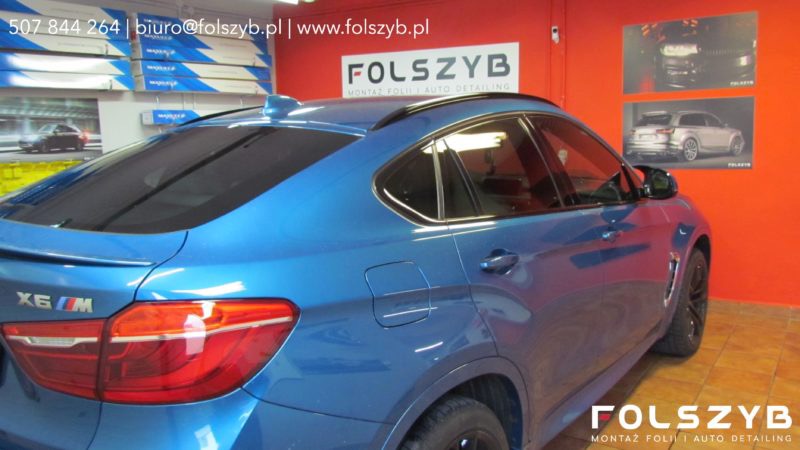 Niebieski samochód BMW X6 M z delikatnie przyciemnionymi szybami, zaparkowany w warsztacie z reklamami firmy Folszyb w tle.