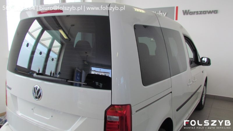 Biały Volkswagen Caddy z przyciemnionymi szybami bocznymi i tylną, zaparkowany w jasnym pomieszczeniu, widok z tyłu i boku.