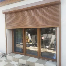 Brązowa roleta zewnętrzna częściowo opuszczona na oknie tarasowym, widok z zewnątrz na taras z meblami ogrodowymi.