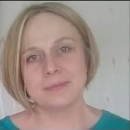 Portret kobiety o jasnych włosach i niebieskich oczach, uśmiechającej się delikatnie do kamery, ubrana w turkusową bluzkę na tle jasnej ściany.