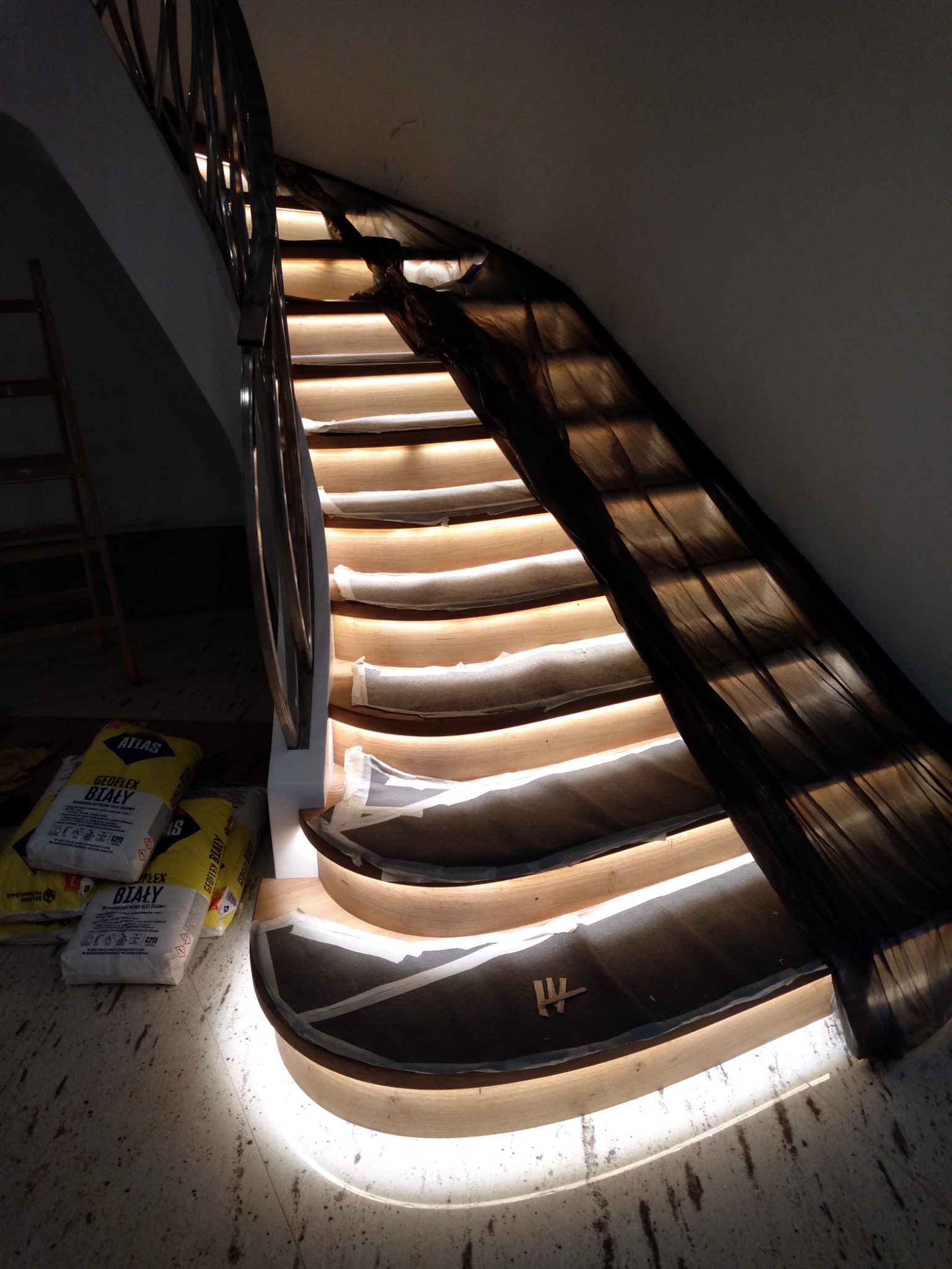 Klatka schodowa z drewnianymi stopniami podświetlonymi ledami, zabezpieczona folią malarską, obok worki z klejem Atlas Geoflex Biały.