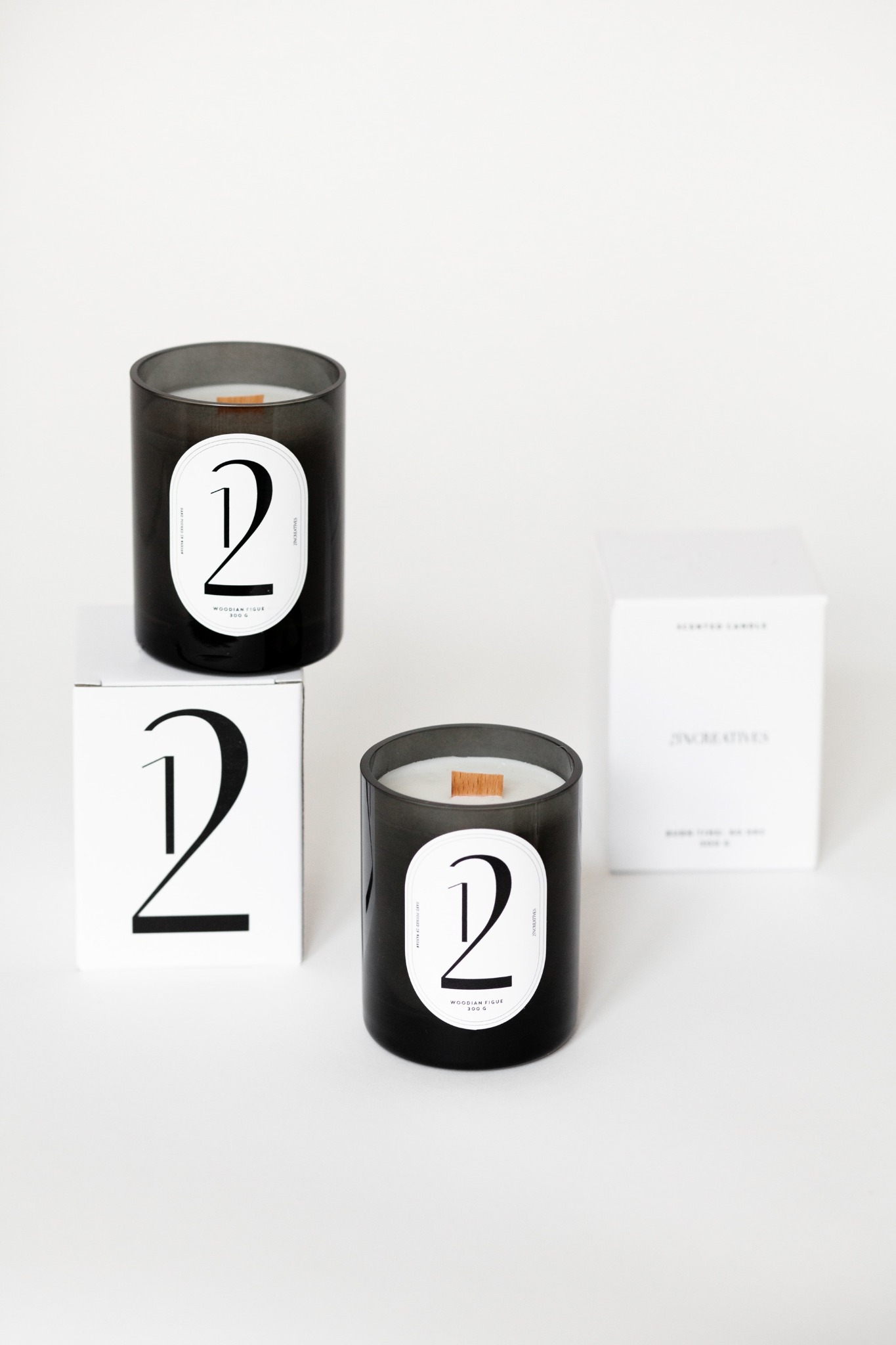 Dwie czarne szklane świece zapachowe z drewnianym knotem i etykietą z cyfrą '2' oraz pudełka na białym tle.