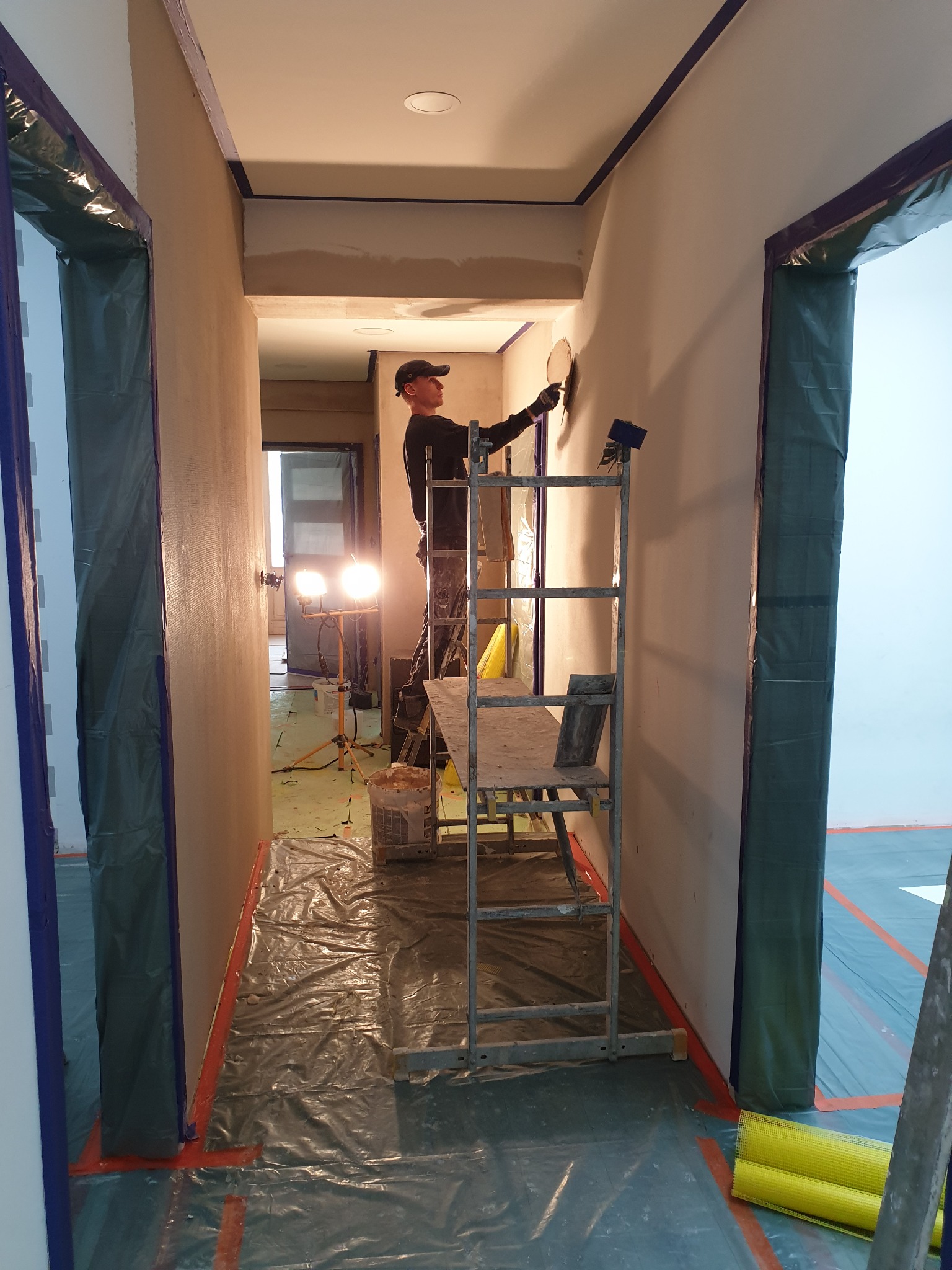 Pracownik na rusztowaniu wyrównuje ścianę w korytarzu, zabezpieczonym folią i taśmą malarską, widoczne lampy budowlane.