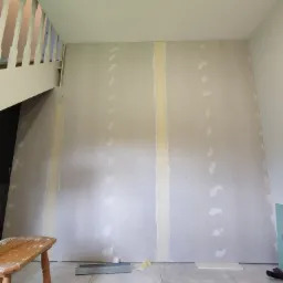 Ściana w trakcie zabudowy płytami gipsowo-kartonowymi w Malczycach, widoczne łączenia wzmocnione taśmą, fragment schodów i drewniany stołek w dolnym lewym rogu.