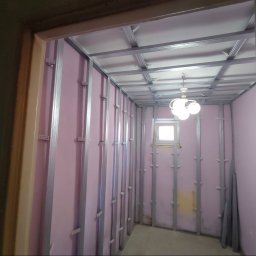 Patryk Stebnicki - Wnętrze w trakcie prac z płytami karton-gips: widoczne metalowe profile, różowa wełna mineralna, okno i lampa. Ujęcie zza futryny drzwi.