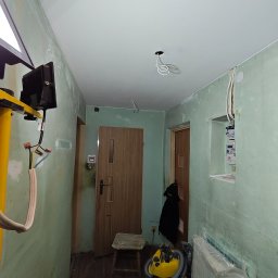 Patryk Stebnicki - Korytarz w trakcie remontu: gładki, biały sufit podwieszany z wystającymi kablami, ściany przygotowane do malowania, widoczny sprzęt budowlany i narzędzia.