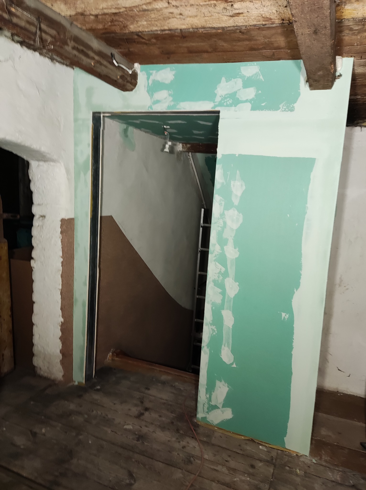 Montaż ościeżnicy z obudową z zielonej płyty gipsowo-kartonowej w surowym wnętrzu, widoczne drewniane belki stropowe i fragment drabiny w przejściu.