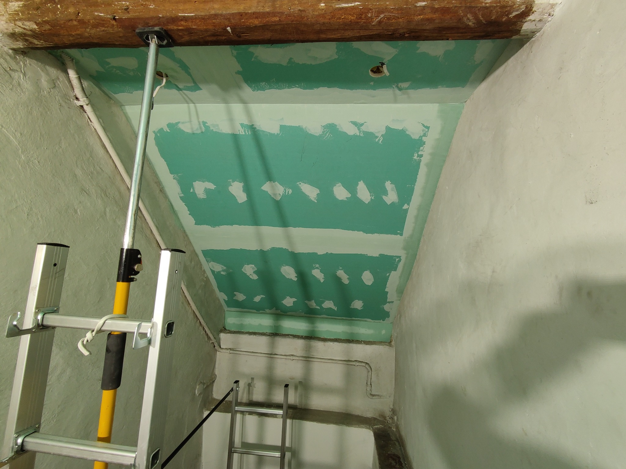 Montaż sufitu z zielonej płyty, widoczne szpachlowane łączenia, podpory teleskopowe i drabina aluminiowa