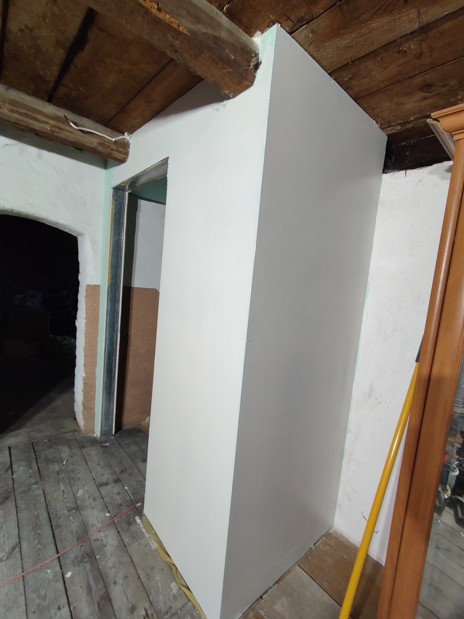 Konstrukcja z profili i białych płyt na poddaszu z drewnianym stropem, widoczne stare deski podłogowe i fragment ościeżnicy drzwiowej.