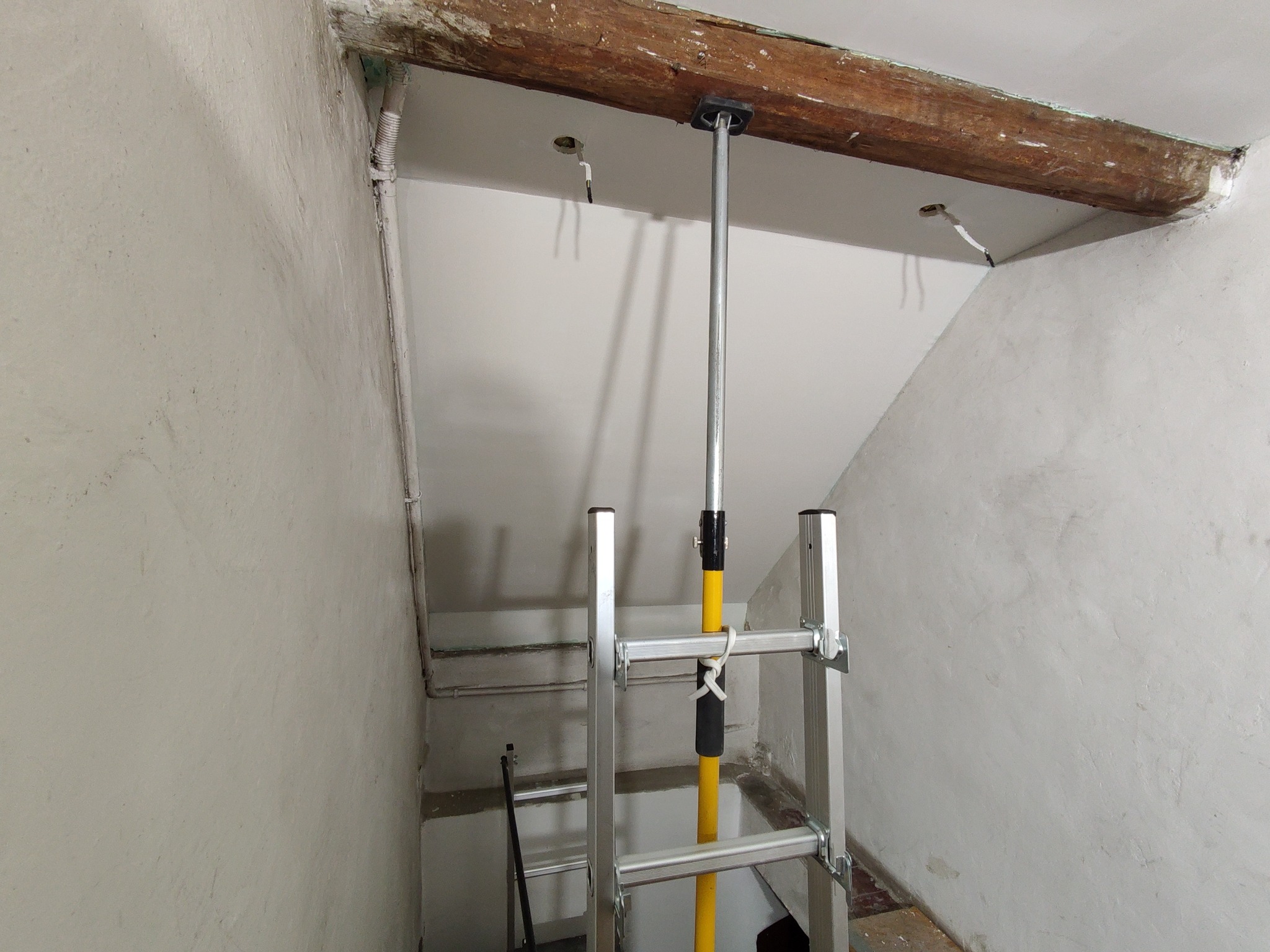 Montaż płyty karton gips na suficie poddasza, widoczna drabina, podpórka teleskopowa i drewniana belka stropowa