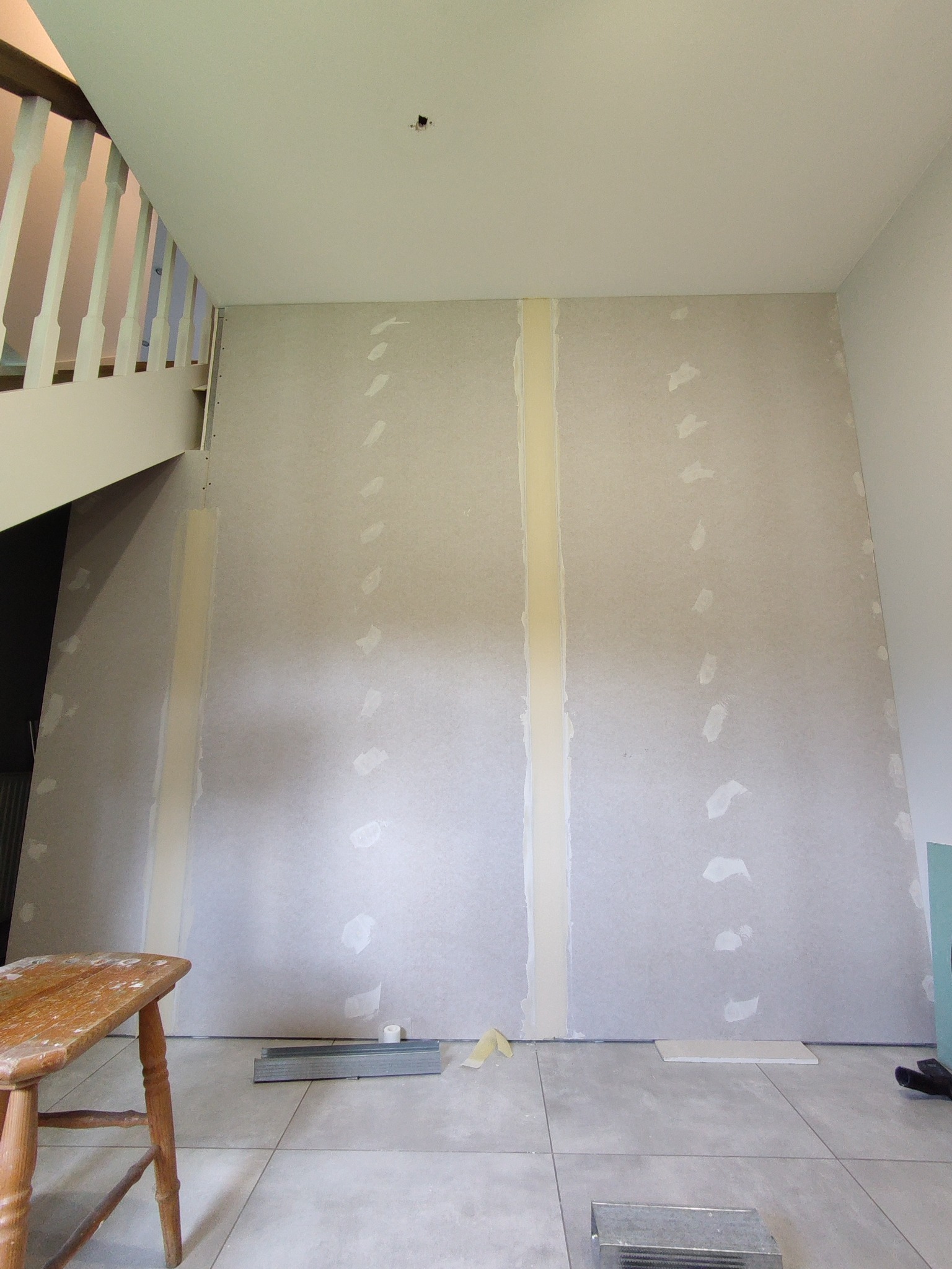 Ściana w trakcie zabudowy płytami gipsowo-kartonowymi w Malczycach, widoczne łączenia wzmocnione taśmą, fragment schodów i drewniany stołek w dolnym lewym rogu.