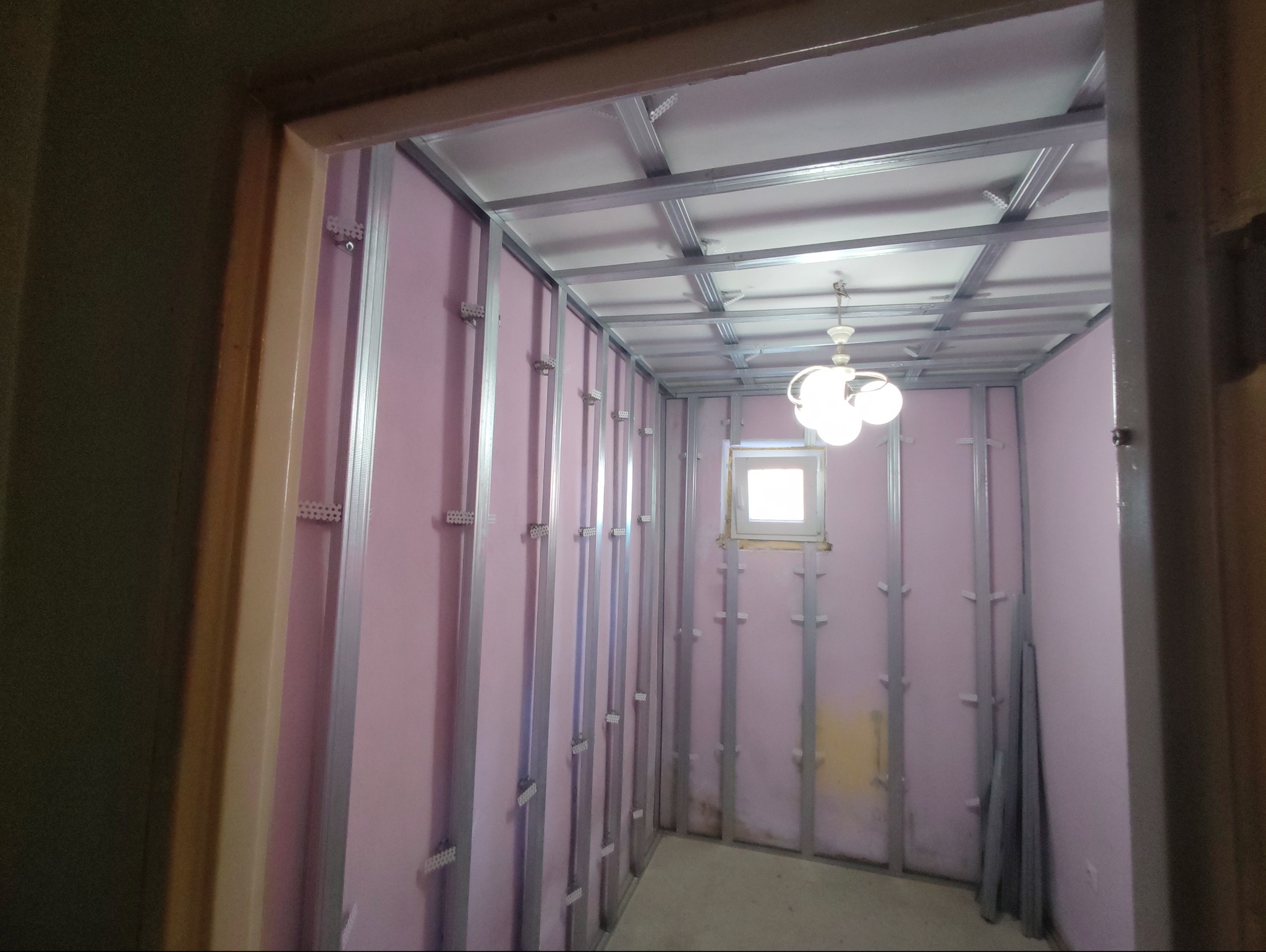 Wnętrze w trakcie prac z płytami karton-gips: widoczne metalowe profile, różowa wełna mineralna, okno i lampa. Ujęcie zza futryny drzwi.