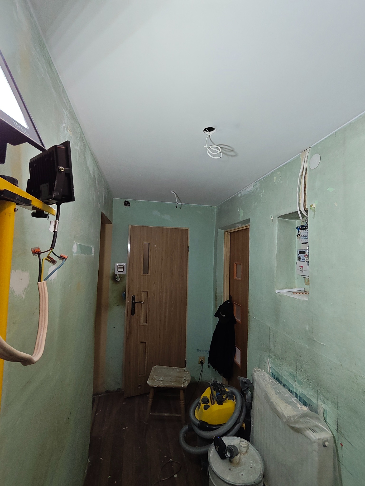 Korytarz w trakcie remontu: gładki, biały sufit podwieszany z wystającymi kablami, ściany przygotowane do malowania, widoczny sprzęt budowlany i narzędzia.