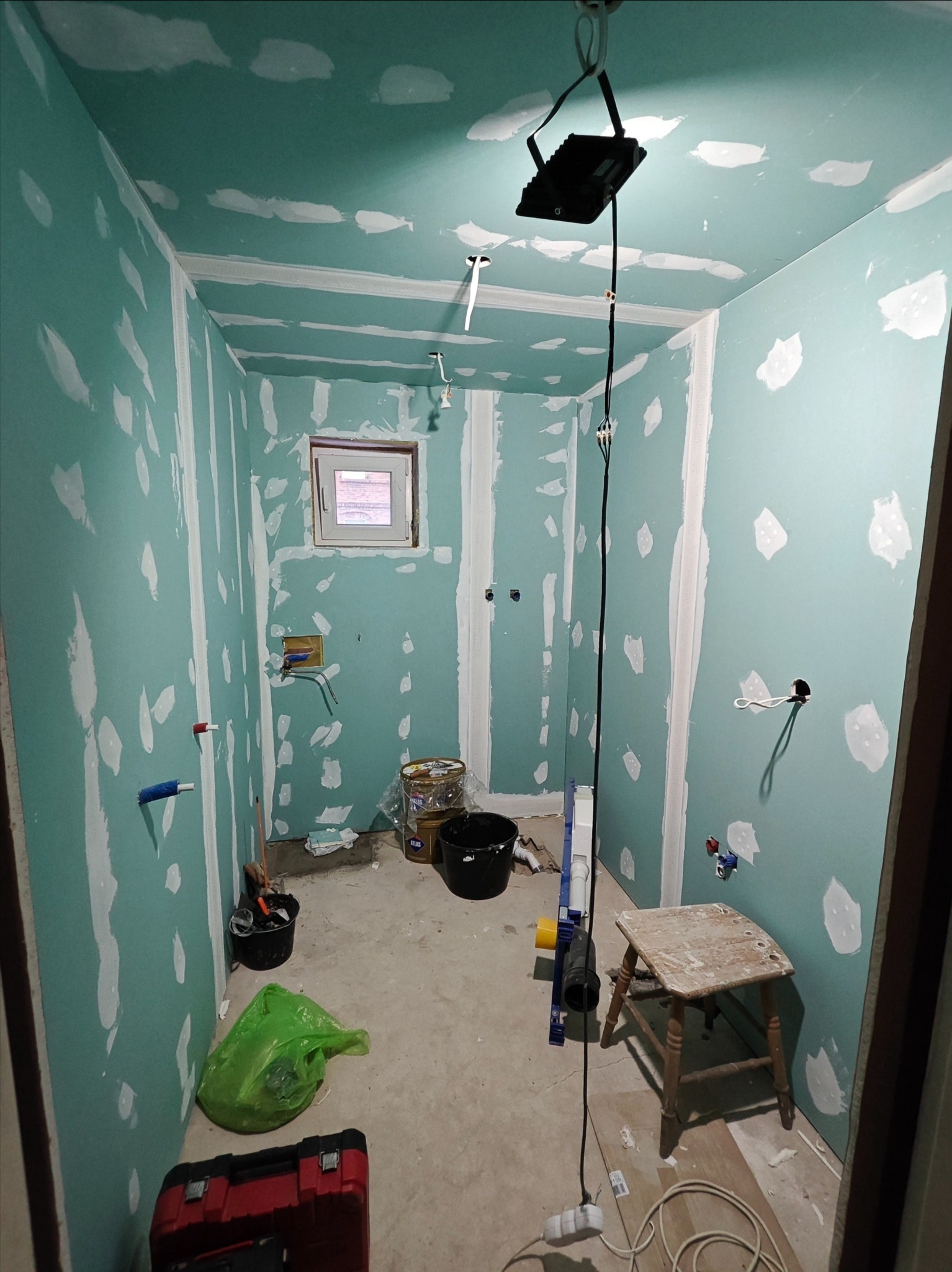Wnętrze w trakcie prac: ściany z płyt gipsowo-kartonowych, widoczne łączenia i szpachlowane miejsca. Na podłodze narzędzia, wiadro i stołek. Okno w tle.