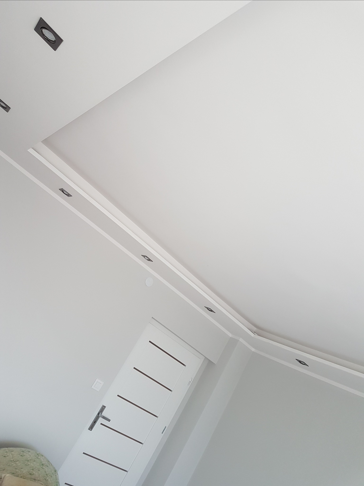 Biały sufit podwieszany z listwami dekoracyjnymi i wbudowanymi punktowymi lampami, fragment białej ściany i drzwi z poziomymi wgłębieniami, widok z dołu.