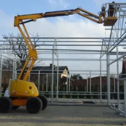 Żółty podnośnik koszowy na budowie szkieletu hali stalowej w Nowym Mieście Lubawskim, widoczne połączenia spawane konstrukcji.