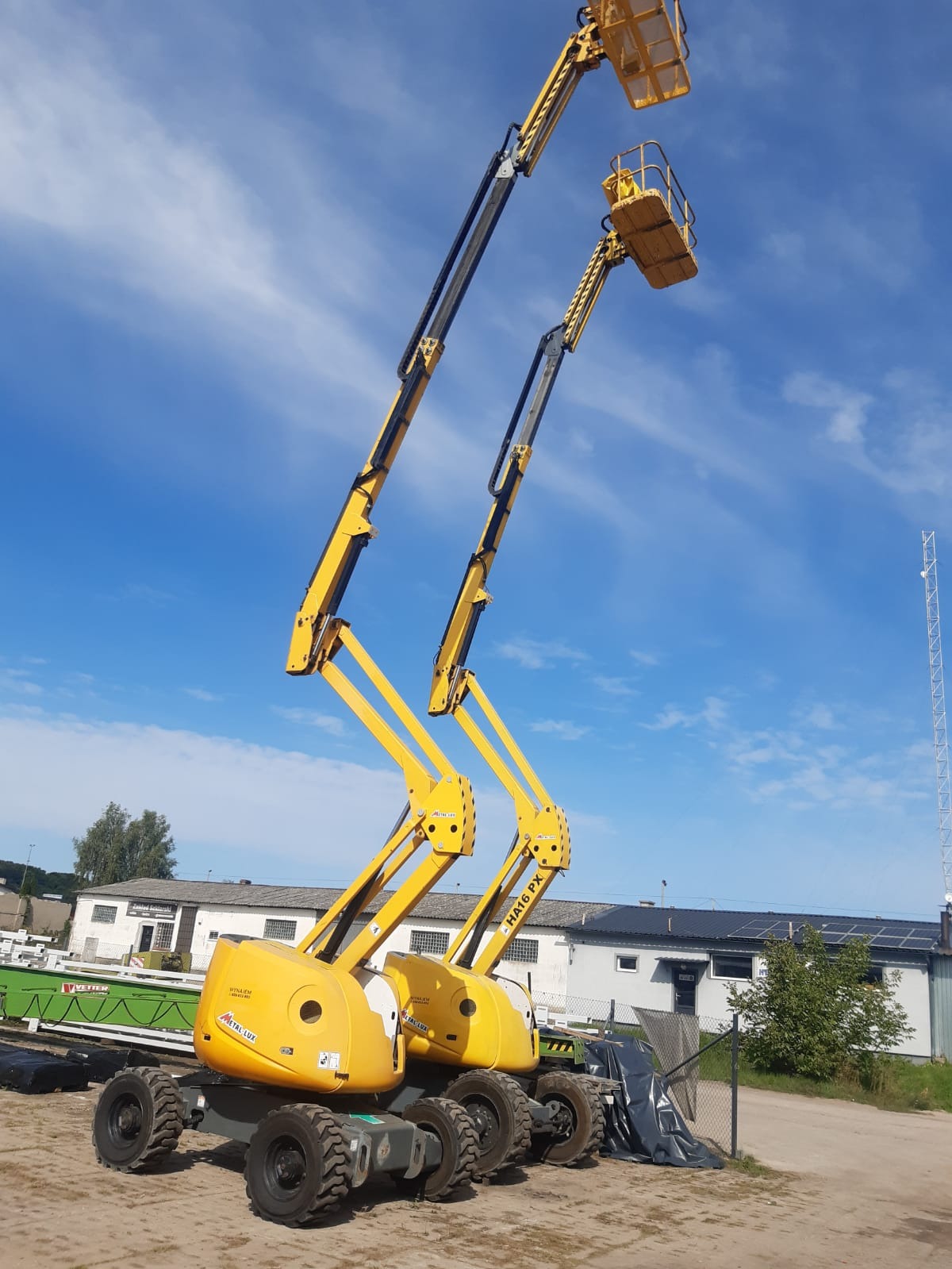 Dwie żółte podnośniki koszowe HA16 PX na nierównym terenie, z wysuniętymi ramionami pod błękitnym niebem z chmurami, w tle budynki z panelami słonecznymi i wieża telekomunikacyjna.