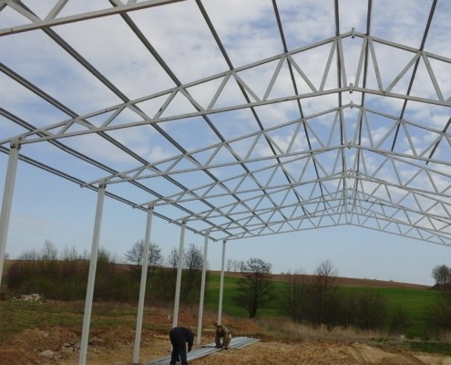 Wznoszenie konstrukcji stalowej hali na tle zielonych pól i błękitnego nieba; widoczni pracownicy montujący elementy na ziemi.