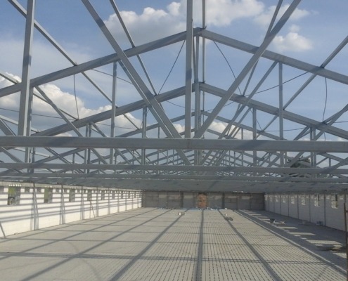 Wnętrze hali w trakcie budowy z widoczną stalową konstrukcją dachu i ścianami z płyt warstwowych, oświetlonych naturalnym światłem wpadającym przez otwory okienne.