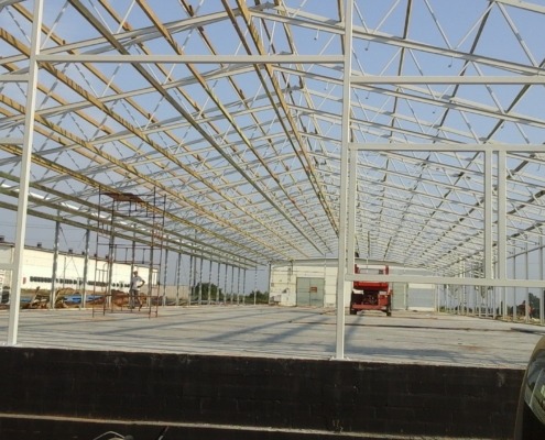 Wnętrze hali w trakcie budowy z widoczną stalową konstrukcją szkieletową, podnośnikiem koszowym i budynkiem w tle.