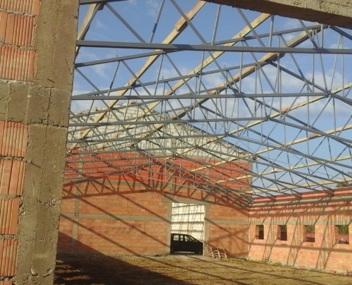 Wnętrze hali w trakcie budowy, widoczne ceglane ściany, stalowa konstrukcja dachu i częściowo zamontowane drewniane elementy więźby dachowej, w tle błękitne niebo.