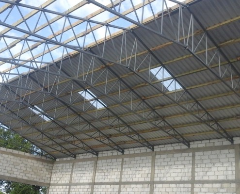 Wnętrze hali w trakcie budowy: widoczna stalowa konstrukcja dachu z drewnianymi belkami i częściowo położonym szarym pokryciem dachowym, ściany z białych bloczków betonowych.