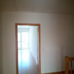 Wnętrze nowo wykończonego mieszkania z drewnianą podłogą, widok na balkon przez otwarte drzwi, klapa rewizyjna na suficie.