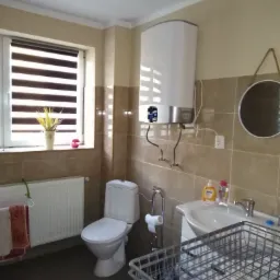 Jasna łazienka z beżowymi płytkami, widoczny bojler elektryczny, toaleta, umywalka z baterią i okrągłe lustro na ścianie. Na parapecie okna dekoracja z suszonych kwiatów w białym wazonie.