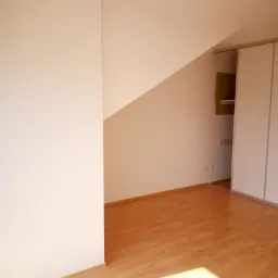 Poddasze z nową, jasną podłogą z paneli imitujących drewno, białe ściany i wbudowana szafa z przesuwnymi drzwiami.