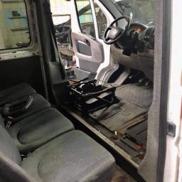 ZaCLEANacz Żdzary - Wnętrze furgonetki w trakcie demontażu, widoczne wymontowane elementy deski rozdzielczej i podłogi, szare fotele z pomarańczowymi zatrzaskami, otwarte drzwi kabiny.