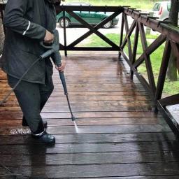 ZaCLEANacz Żdzary - Mężczyzna w ciemnej kurtce i gumowych butach czyści drewniany taras za pomocą myjki ciśnieniowej, widoczny efekt czyszczenia i brudna woda.